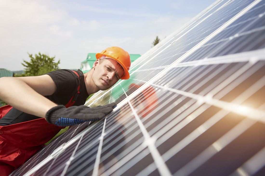 A solar installer wearing an orange hard hat and red overalls inspects solar panel angle