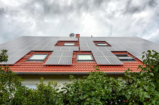 A house’s roof covered in solar panels in front of green shrubs on a cloudy day