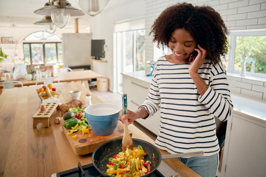 A smiling woman talks on the phone while cooking a healthy meal in her brightly lit kitchen