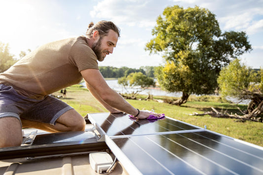 An adventurer wearing a t-shirt and jean shorts installs an off-grid solar system on a trailer in a grassy area