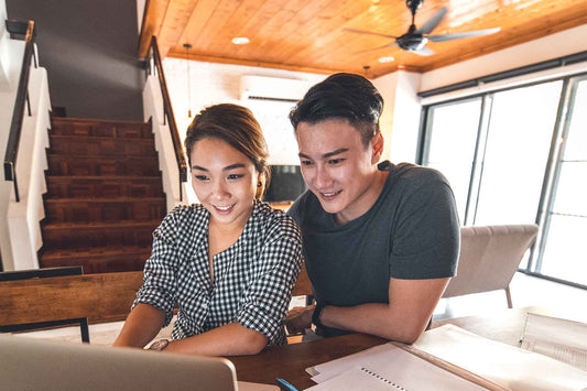 A smiling couple sitting at their dining room table in front of a laptop and notebooks