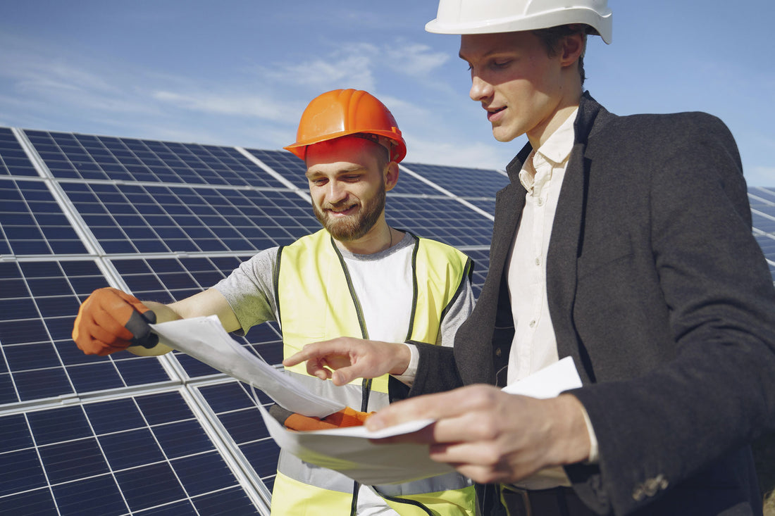 A solar installer wearing a yellow vest and a man wearing a suit discuss documents in front of a solar panel array